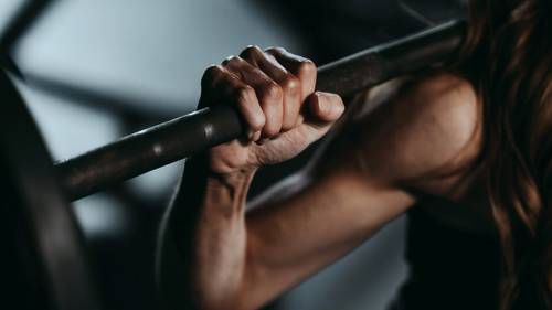 Close-up of a person gripping a barbell, highlighting a muscular arm and hand, set against a blurred gym background.