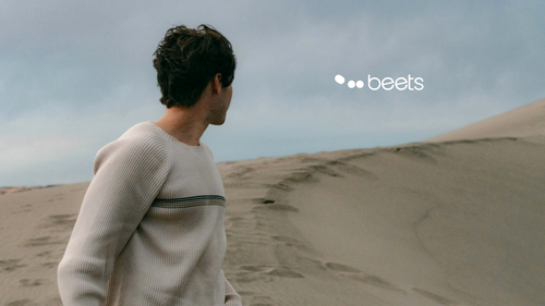 Eine Person in einem leichten Pullover steht unter bewölktem Himmel auf Sanddünen und schaut zur Seite. In der oberen rechten Ecke steht das Wort „beets“.
