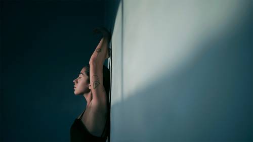 Frau mit Tattoos streckt sich an einer Wand, die Augen geschlossen, im sanften Licht.