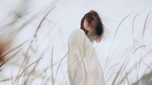 Eine Frau in einem weißen Pullover steht im Freien, mit vom Wind verwehten Haaren und hohem Gras im Vordergrund vor einem wolkigen Himmel.