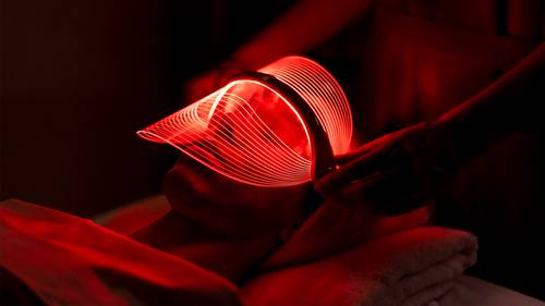 Person, die in einem schwach beleuchteten Raum eine LED-Lichttherapie mit einer leuchtend roten Maske auf dem Gesicht erhält.