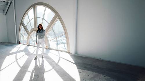 Woman in a gray hoodie and white pants stands in a sunlit room with large, arched windows casting shadows on the floor.