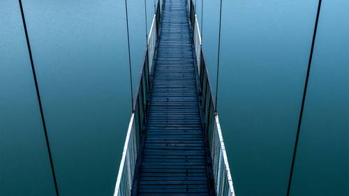 Draufsicht auf eine schmale Hängebrücke mit Holzplanken und weißen Geländern, die sich über ruhiges blaues Wasser erstreckt.