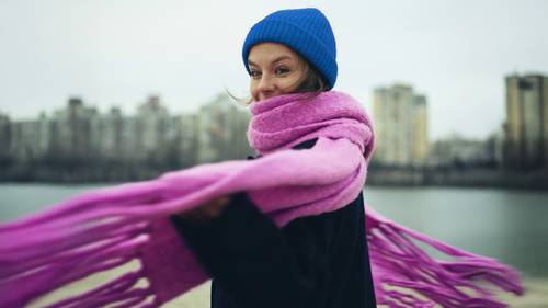 Una persona con gorro azul y bufanda rosa sonríe, de pie cerca de un cuerpo de agua con edificios de la ciudad al fondo.