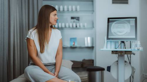 O femeie stă într-o clinică modernă și privește un ecran de computer cu informații medicale, cu rafturi de produse în fundal.