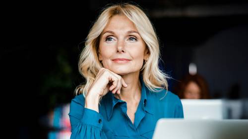 Blonde Frau in einem blauen Hemd, die drinnen sitzt, ihr Kinn auf die Hand stützt und nachdenklich zur Seite schaut.
