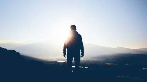 Die Silhouette einer Person, die auf einem Hügel steht, während die Sonne über den fernen Bergen aufgeht und einen hellen Schein sowie Schatten in der Umgebung wirft.