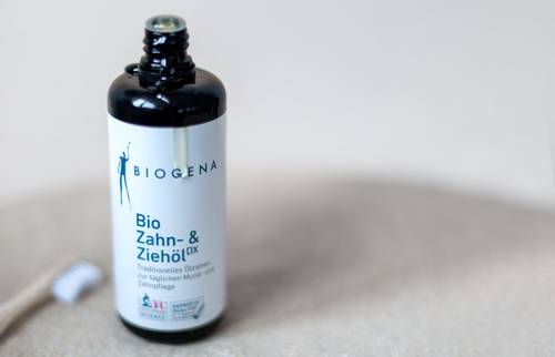 Eine Flasche BIOGENA Bio Zahn- & Ziehölox Öl auf einer hellen Oberfläche, mit einer unscharfen Zahnbürste im Hintergrund.