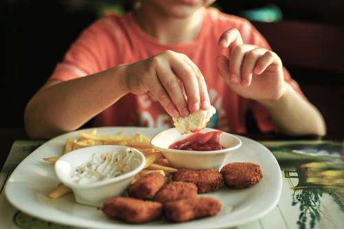 Child dipping a chicken nugget into ketchup on a plate with fries and another sauce.