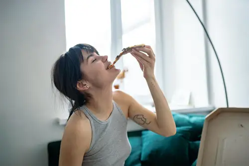 Eine Person in einem grauen Tanktop sitzt auf einem blaugrünen Sofa in der Nähe eines Fensters und isst genüsslich ein Stück Pizza.