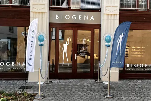 Ladenfront von Biogena mit Glastüren, flankiert von zwei Markenflaggen und Ballondekorationen, auf einem gepflasterten Gehweg.