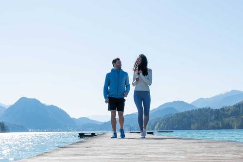 Un hombre y una mujer vestidos con ropa deportiva pasean por una pasarela de madera junto a un pintoresco lago, con las montañas y un cielo despejado al fondo.