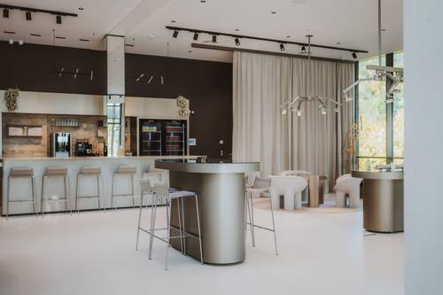 Moderner Barbereich mit eleganten Barhockern, minimalistischen Tischen und zeitgemäßer Beleuchtung. Große Fenster und neutrale Farbtöne schaffen eine luftige Atmosphäre.