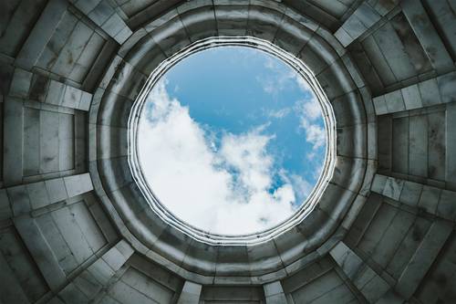 Blick auf einen blauen Himmel mit Wolken durch eine runde Steinöffnung, die einem Oberlicht ähnelt und von konzentrischen architektonischen Schichten umgeben ist