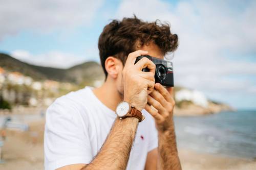 Persoană într-o cămașă albă fotografiind cu o cameră pe o plajă, purtând un ceas, cu munți și ocean încețoșați în fundal.