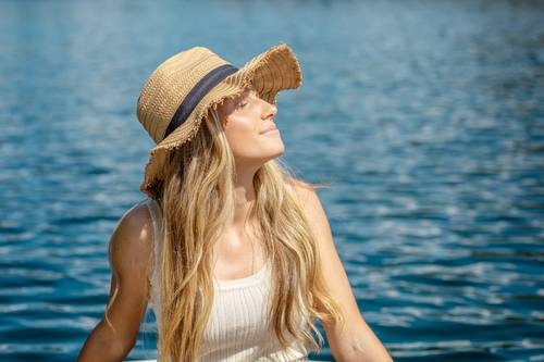 Eine Frau mit Strohhut und weißem Tanktop genießt die Sonne an einem blauen See und blickt mit einem gelassenen Ausdruck nach oben.