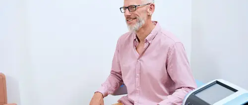 Ein Mann mit Brille und Bart, der ein rosa Hemd trägt, sitzt lächelnd in einem hellen Raum in der Nähe von medizinischem Equipment.