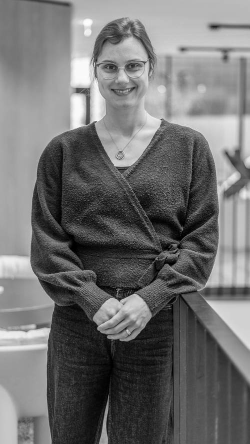 Lächelnde Person mit Brille, bekleidet mit einem dunklen Pullover und Jeans, steht in einem Innenraum vor einem unscharfen Hintergrund. Schwarz-Weiß-Foto.