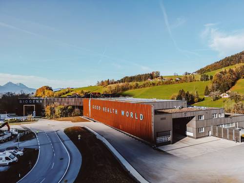 Ein modernes Industriegebäude mit der Aufschrift „Biogena Good Health World“, umgeben von üppig grünen Hügeln und einem strahlend blauen Himmel.