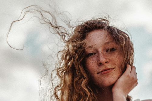 Close-up of a person with curly hair and freckles, smiling softly against a cloudy sky background.