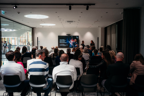 Audience in a conference room facing a large screen displaying a presentation.