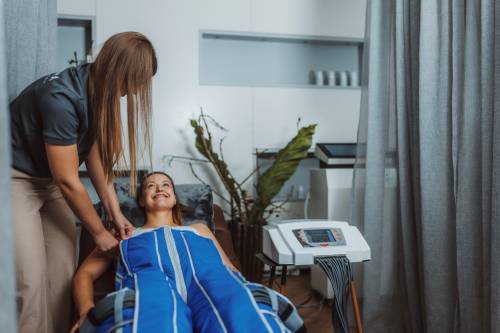 Eine Frau liegt auf einem Spa-Bett und trägt einen blauen Kompressionsanzug. Sie lächelt eine Begleiterin an, die den Anzug in einem ruhigen Raum anpasst.