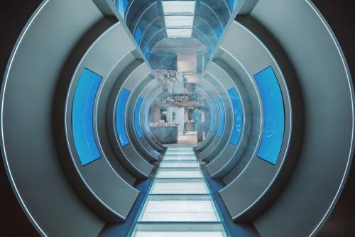 Futuristischer Flur mit kreisförmigem Design, beleuchtet von blauen Lichtern und gesäumt von Fenstern, der zu einem hellen, offenen Raum führt.