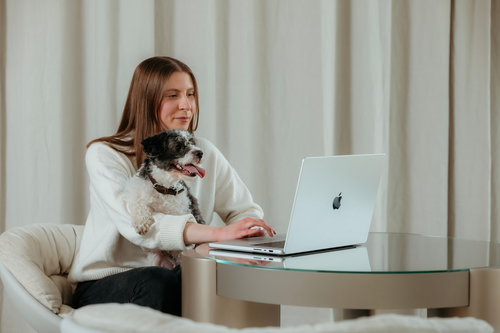 A woman in a white sweater sits at a table using a laptop with a small black and white dog on her lap.