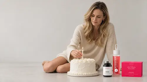 Woman in a beige sweater lighting a candle on a white cake, sitting next to skincare products and a collage powder package on the floor.