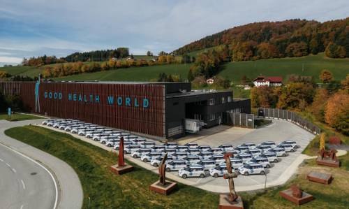 Luftaufnahme eines großen Gebäudes mit der Aufschrift „GOOD HEALTH WORLD“, vor dem zahlreiche weiße Autos geparkt sind, umgeben von herbstlichen Bäumen und Hügeln.