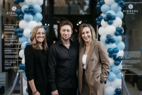Three people smiling in front of a building entrance, decorated with blue and white balloons.