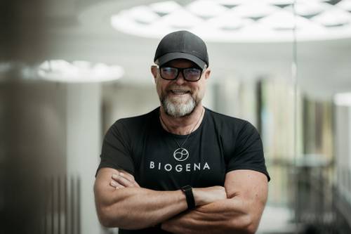 Ein Mann in einem schwarzen BIOGENA-Shirt und einer Kappe, der mit verschränkten Armen lächelt und in einem Innenraum unter einem hellen, runden Licht steht.