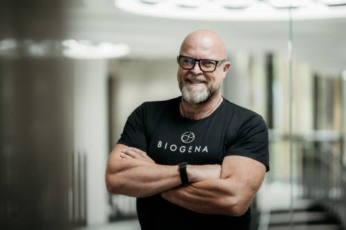 Glatzköpfiger Mann mit Brille und Bart, der ein schwarzes BIOGENA-Shirt trägt, lächelt mit verschränkten Armen in einem modernen, hellen Innenraum.