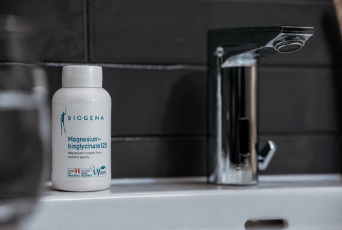 A bottle of Biogena Magnesium-bisglycinate 125 on a white counter beside a shiny faucet, against a dark tiled wall.