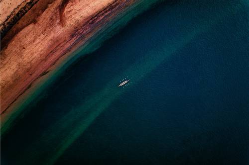 Vedere aeriană a unei bărci solitare cu vâslași pe o mare vastă, de un albastru profund, în apropierea unei coaste nisipoase.