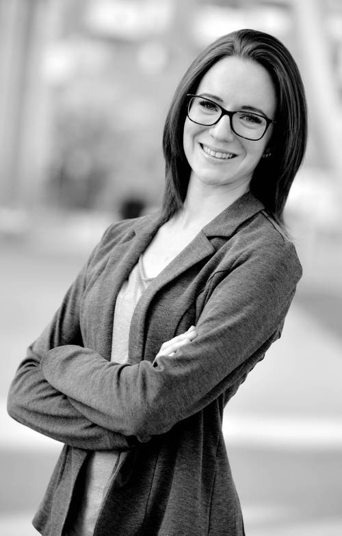 Lächelnde Frau mit Brille und dunklem Haar, die einen Blazer trägt, steht mit verschränkten Armen in einer unscharfen Außenumgebung. Schwarz-Weiß-Foto.