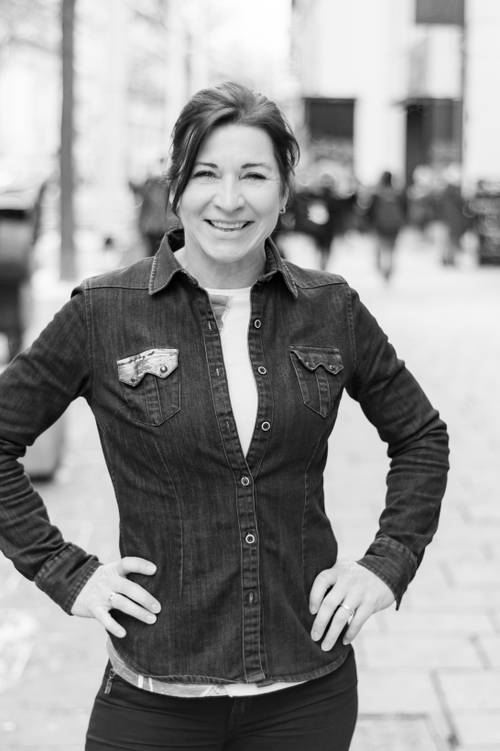 Lächelnde Frau in einer Jeansjacke steht selbstbewusst mit den Händen in den Hüften auf einer belebten Straße in der Stadt, Schwarz-Weiß-Foto.