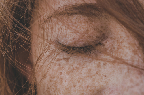 Nahaufnahme eines geschlossenen Auges mit langen Wimpern, umgeben von Sommersprossen und roten Haarsträhnen.
