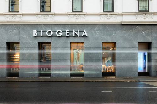Street view of BIOGENA store with grey marble facade and large windows. Blurred lights from passing traffic suggest motion.
