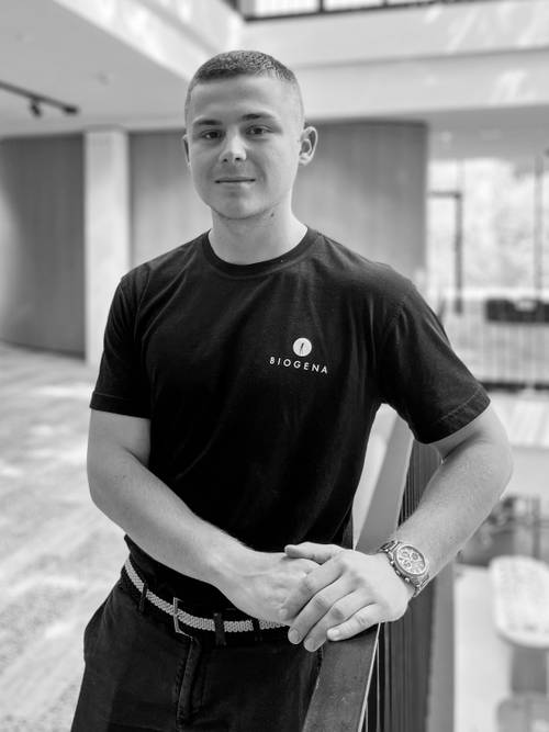 Mann in einem schwarzen BIOGENA-T-Shirt steht in einem Innenraum und lehnt sich an ein Geländer. Schwarz-Weiß-Foto mit natürlicher Beleuchtung und moderner Inneneinrichtung.
