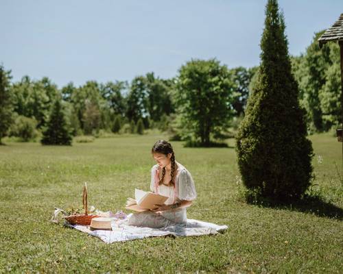O femeie cu părul împletit stă pe o pătură într-o poiană însorită, citind o carte. Un coș de picnic se află lângă ea, înconjurat de copaci.