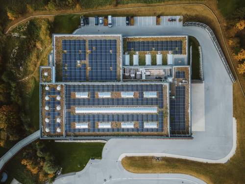 Vedere aeriană a unei clădiri cu panouri solare pe acoperiș, înconjurată de o parcare și zone verzi.