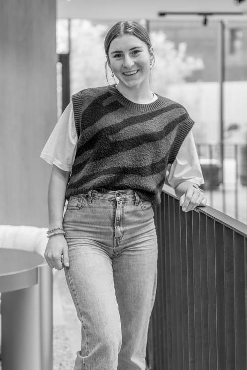 Person lächelt, steht drinnen an einem Geländer, trägt eine gestreifte Strickweste über einem T-Shirt und Jeans. Schwarz-Weiß-Foto.