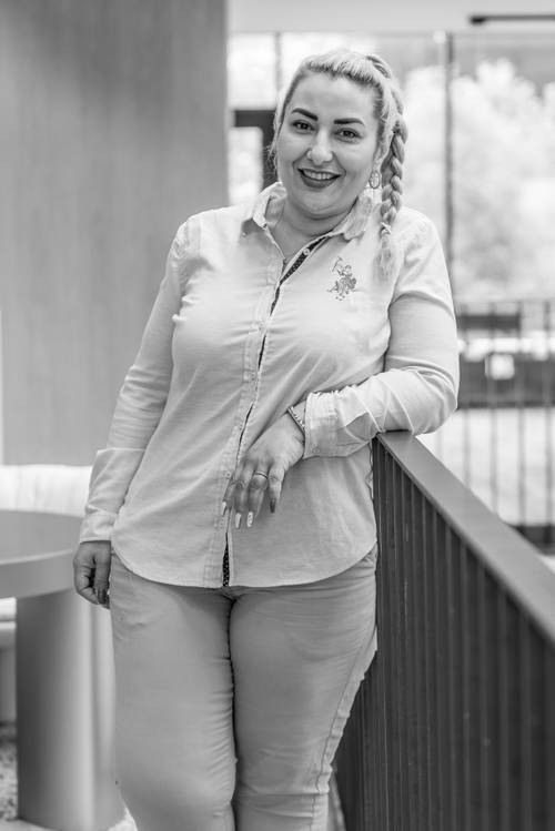 Frau mit geflochtenen Haaren, bekleidet mit einem hellen Hemd und einer Hose, lehnt sich drinnen an ein Geländer und lächelt in die Kamera. Schwarz-Weiß-Foto.