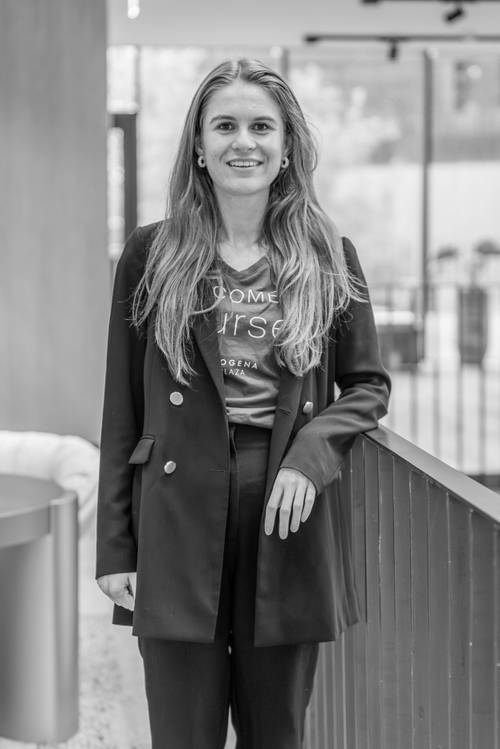 Schwarz-Weiß-Foto einer Frau mit langen Haaren in einem Blazer, die in einem Innenraum steht, sich an ein Geländer lehnt und in die Kamera lächelt.