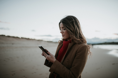 Woman in a red sweater and brown coat using a smartphone on a deserted beach at dusk.