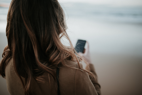 Eine Frau mit braunen Haaren, von hinten gesehen, schaut auf ihr Handy, während sie in der Nähe eines Strandes steht.