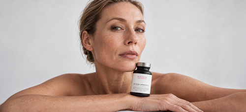 A woman poses with a bottle labeled "Biogena Aesthetics NR Skin Longevity+" resting on her shoulder against a plain background.