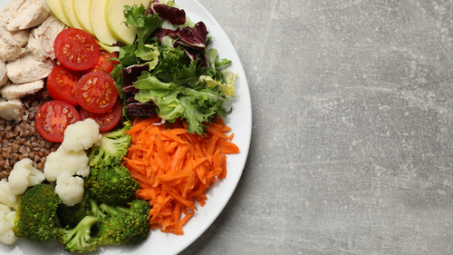 Ein Teller mit Hähnchenscheiben, Tomaten, Äpfeln, gemischtem Blattsalat, geraspelten Möhren, Blumenkohl, Brokkoli und Buchweizen auf einer grauen, strukturierten Unterlage.