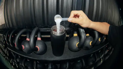 Eine Hand schaufelt Pulver in eine Shaker-Flasche, umgeben von Kettlebells, die auf einem Gestell aufgereiht sind.
